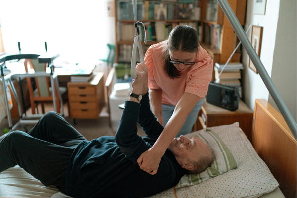Caregiver helps an elderly man out of bed with the use of an assistive lifting device